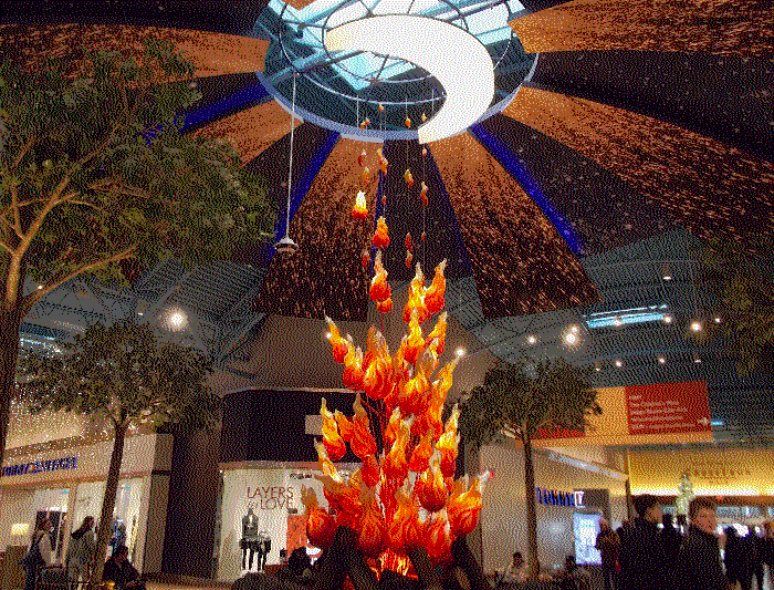 A photo of the ceiling behind the white cowboy, which is a glowing crescent moon with glittering 3d rays. A sculpted bonfire, made up of individual hanging flames, glows and reaches for the moon.