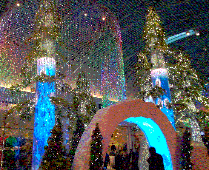 A photo of an intricate christmas display, tall fake trees adorned in screens with glowing ice textures, and rainbow curtain lights hanging from the ceiling in a fake aurora borealis.